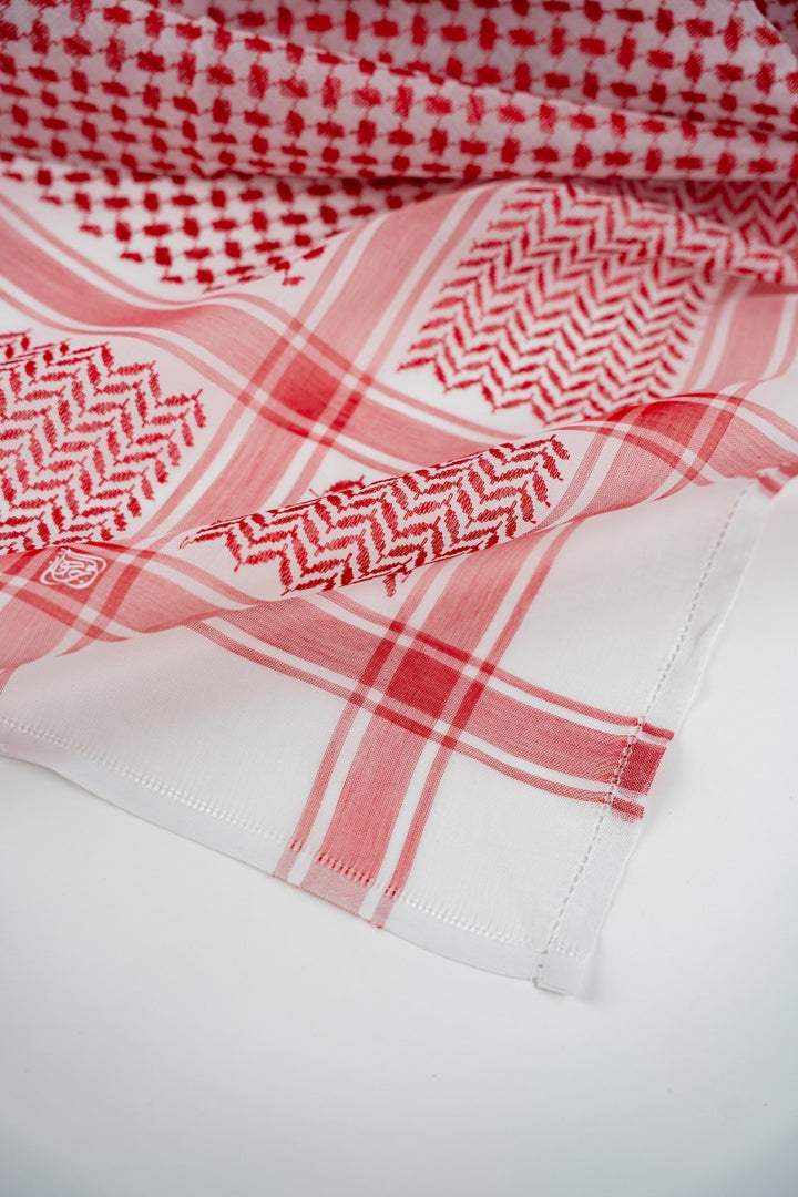 A close-up shot of a red and white men’s shemagh, softly folded on a white surface. The image clearly shows the traditional geometric patterns, red checkered lines, woven texture, and neatly stitched edges.